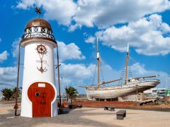 Small,Lighthouse,With,Sailing,Ship,On,Land,In,Harbor,Area